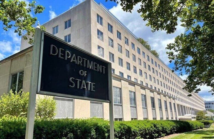 A "Department of State" sign stands in front of a large, multi-story stone office building with rows of windows, surrounded by green trees and hedges.