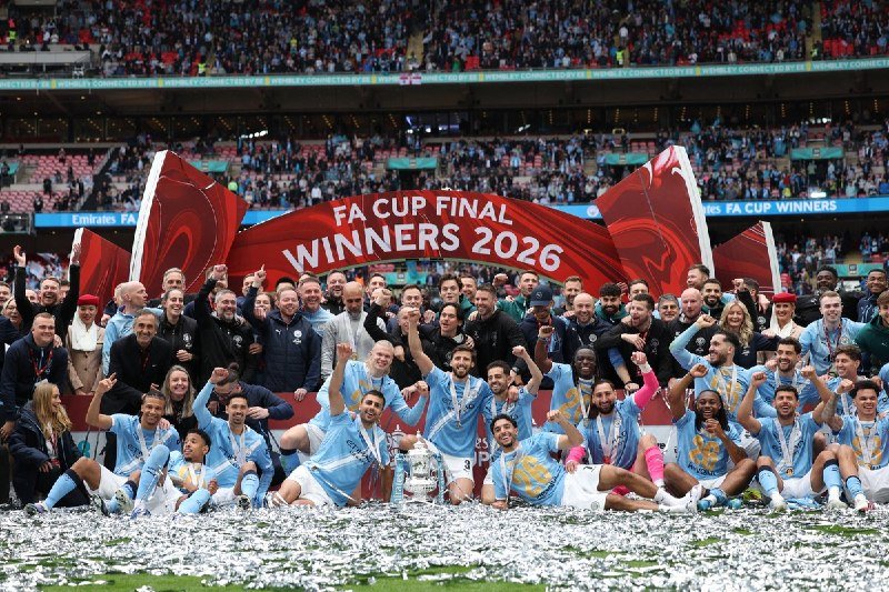 A football team in light blue kits poses with the FA Cup trophy beneath a "Winners 2026" banner amid confetti, surrounded by staff and a packed stadium crowd.