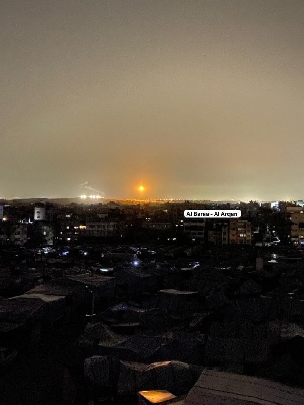 A bright orange flash illuminates the night horizon over a darkened cityscape, with a labeled graphic reading "Al Baraa - Al Arqan" in the sky.