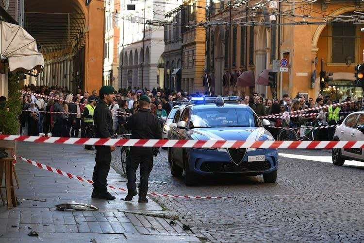 Two uniformed officers stand behind red-and-white police tape near a marked Italian police car on a cobblestone street, as a crowd watches from the background.