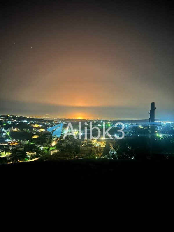 Nighttime cityscape with illuminated buildings and a glowing horizon, featuring a watermark reading "Alibk3."