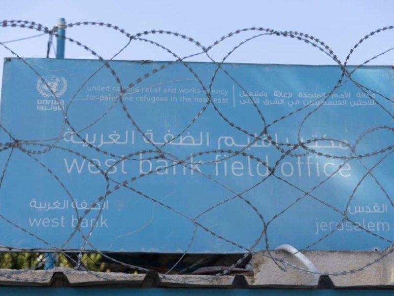 A blue UNRWA sign for the "West Bank Field Office" in Jerusalem is visible behind coiled razor wire atop a wall.