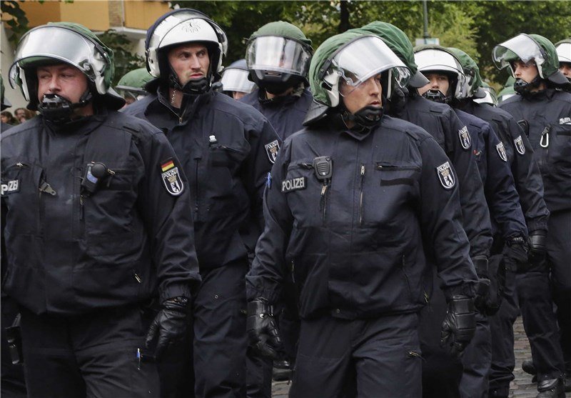 Police officers in dark uniforms and helmets with visors march in formation, their uniforms bearing "POLIZEI" patches.