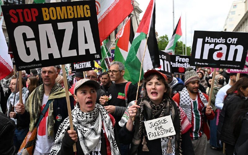 A crowd of demonstrators holding Palestinian flags and protest signs reading "STOP BOMBING GAZA," "Hands Off Iran," and "Justice for Palestine" march through a street.