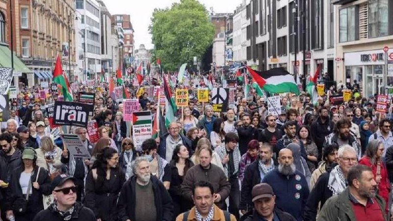 A large crowd marches down a city street carrying Palestinian flags, a peace symbol sign, and placards reading "Hands Off Iran" and "Stop the war."