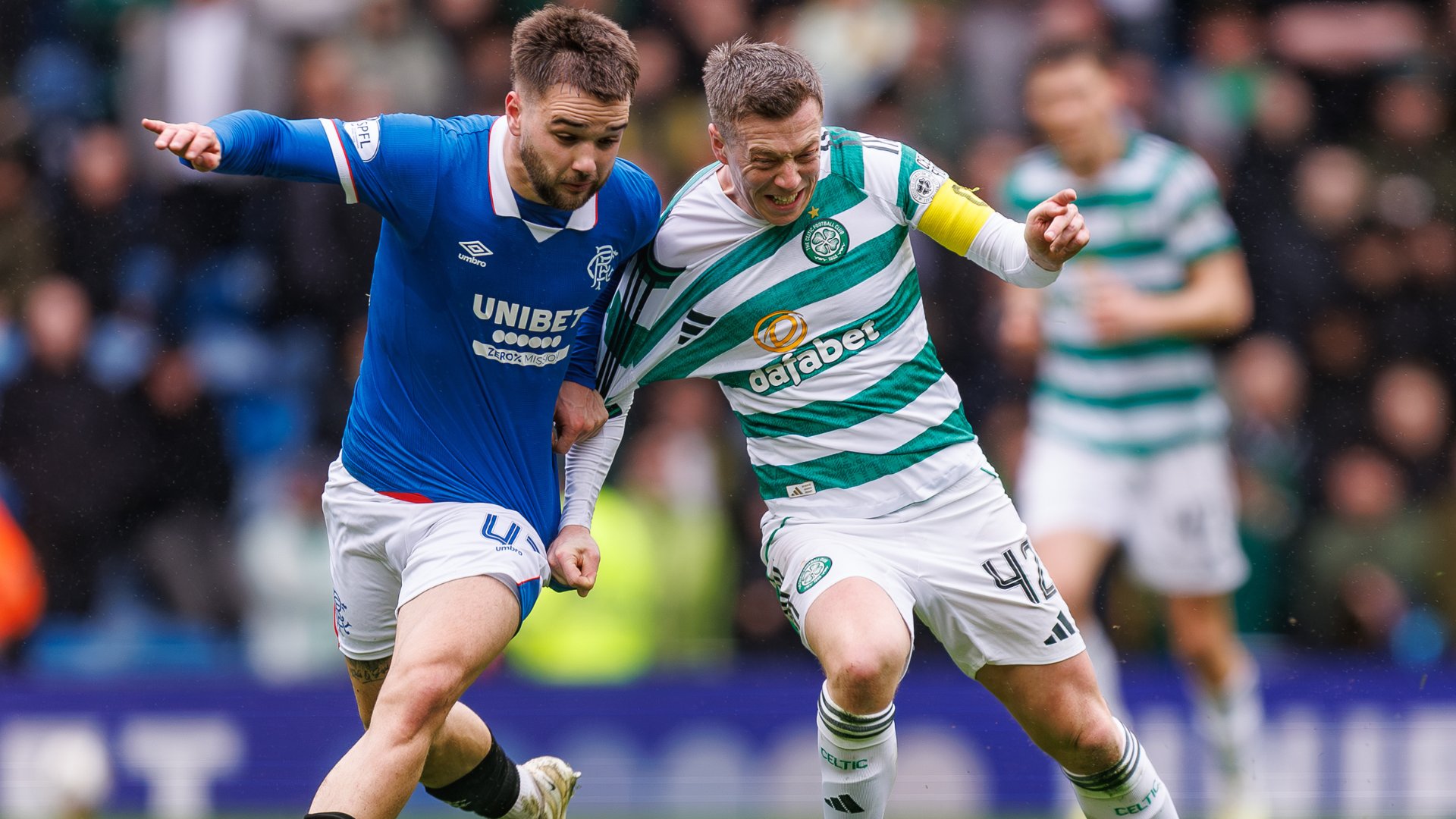 Two soccer players, one in a blue Rangers jersey and the other in a green-and-white hooped Celtic kit, battle for the ball during a match in front of a stadium crowd.