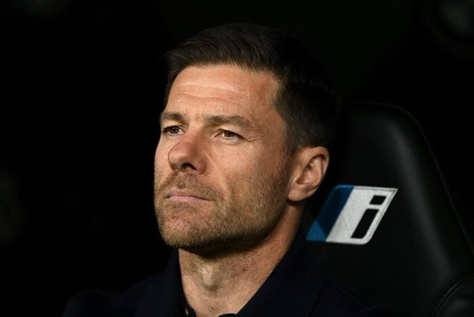A man with short dark hair and a trimmed beard sits in a black chair against a dark background, looking off to the side.