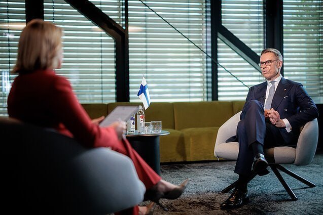 A woman in red holds papers while interviewing a suited man in a modern room with a Finnish flag on the table.