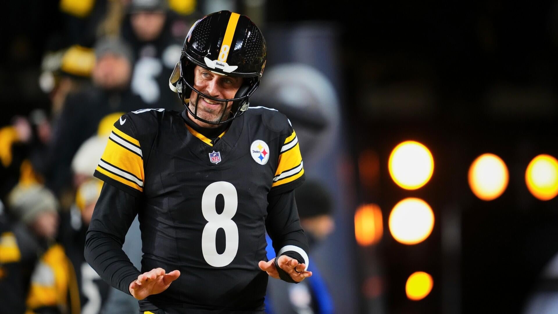 A football player wearing a black Steelers jersey with the number 8 and matching helmet smiles while walking on the sideline, with stadium lights blurred in the background.