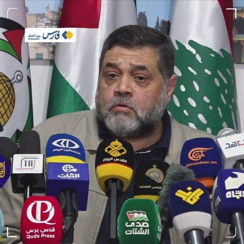A bearded man speaks at a press conference behind a row of microphones labeled with Arabic media logos, with Lebanese flags visible in the background.