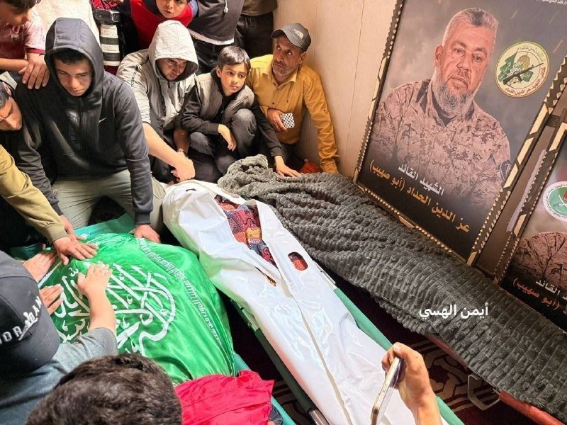 Mourners gather around a body wrapped in white and green cloth, with a large framed portrait of a bearded man and Arabic text displayed nearby.
