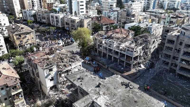 An aerial view shows extensive destruction in an urban neighborhood, with several multi-story buildings reduced to rubble and others showing severe structural damage, while crowds gather in the streets below.
