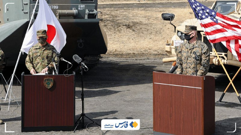 Two masked military officers stand at separate podiums outdoors, flanked by a Japanese flag and an American flag, with military vehicles positioned behind them on a grassy field.