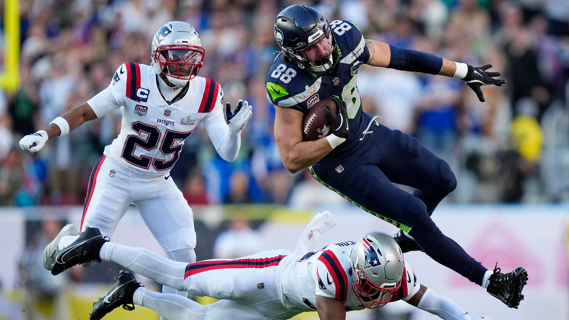 A Seattle Seahawks tight end leaps over a fallen New England Patriots defender while carrying the football, as another Patriots player gives chase during an NFL game.