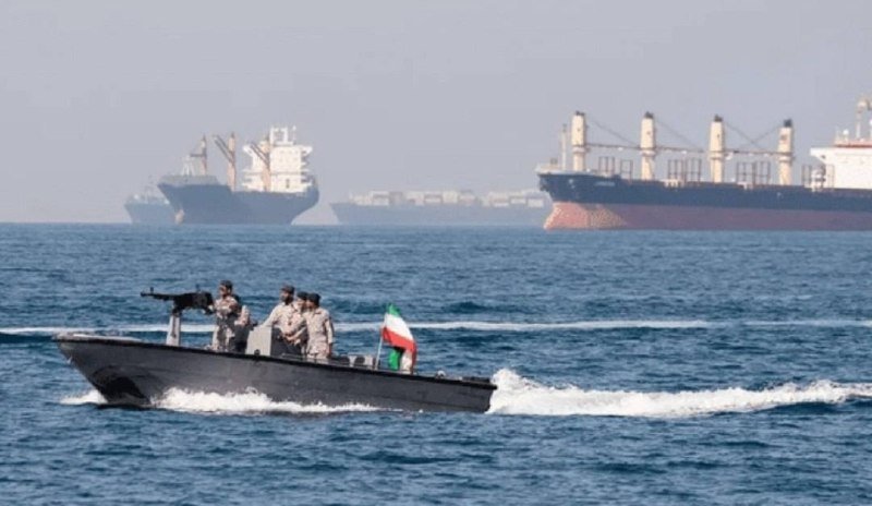 Armed personnel in a small patrol boat flying an Iranian flag speed across the sea, with large cargo ships visible in the hazy background.