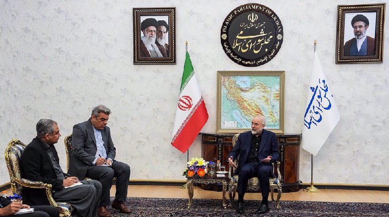 Four men sit in ornate chairs during a meeting in a room displaying framed portraits, an Iranian flag, a map, and an emblem reading "Islamic Parliament of Iran."