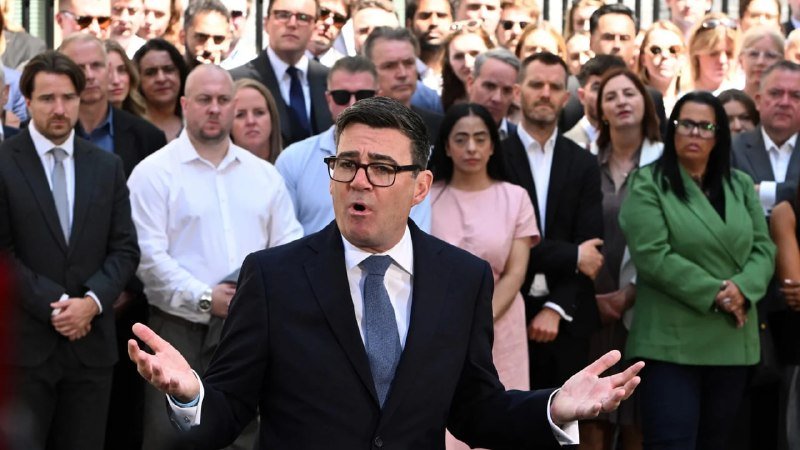 A bespectacled man in a dark suit and blue tie speaks with outstretched arms, standing before a large group of attentive onlookers gathered outdoors.