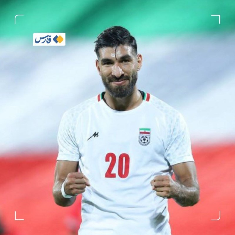 A bearded soccer player wearing a white Iran jersey with the number 20 clenches both fists in a celebratory gesture.