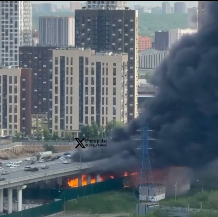 Thick black smoke billows from a burning structure beneath an elevated highway in an urban area, with cars traveling on the road and high-rise buildings visible in the background.