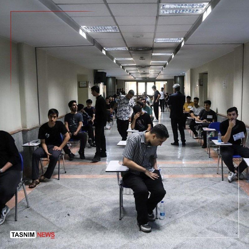 Young men sit at small desks lined along both sides of a long, fluorescent-lit hallway, appearing to take an exam or test.