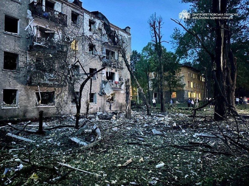 A heavily damaged multi-story apartment building with shattered windows and debris scattered across the ground, with several people standing nearby amid broken tree branches.