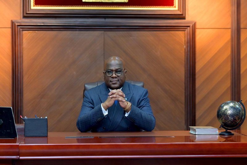 A man in a blue suit and glasses sits at a wooden desk with hands clasped, beside a laptop, book, and globe, against a wood-paneled wall.