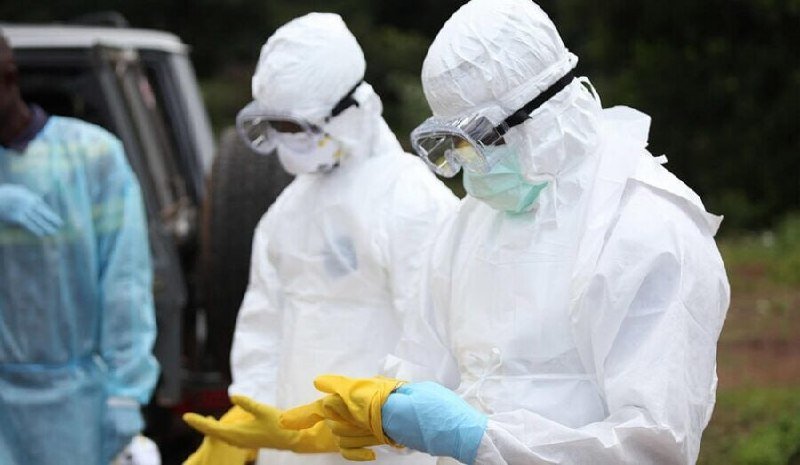 Two people in white hazmat suits, goggles, and face masks stand outdoors while one adjusts yellow gloves, with a third person in a blue gown visible nearby.