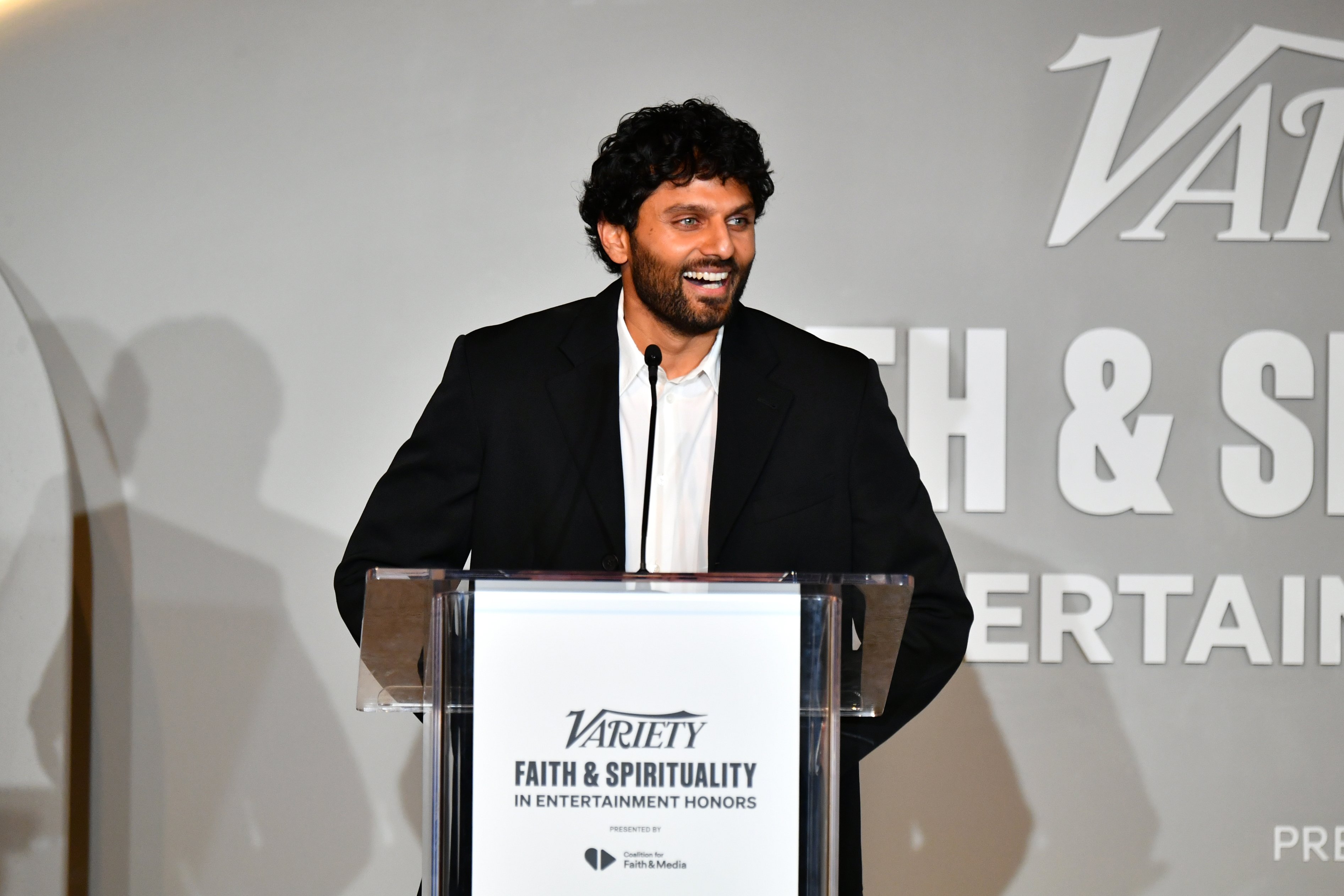 A smiling man in a black suit speaks at a clear podium displaying the "Variety Faith & Spirituality in Entertainment Honors" logo during an awards event.