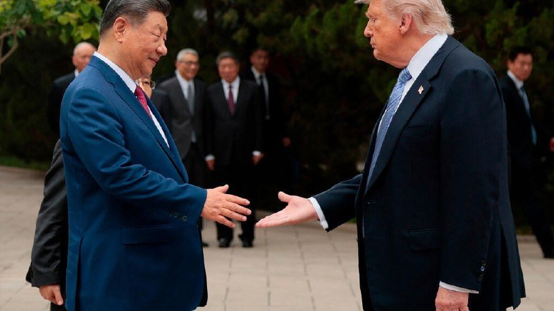 Two men in dark suits extend their hands toward each other outdoors on a paved surface, with several other suited men standing in the background.