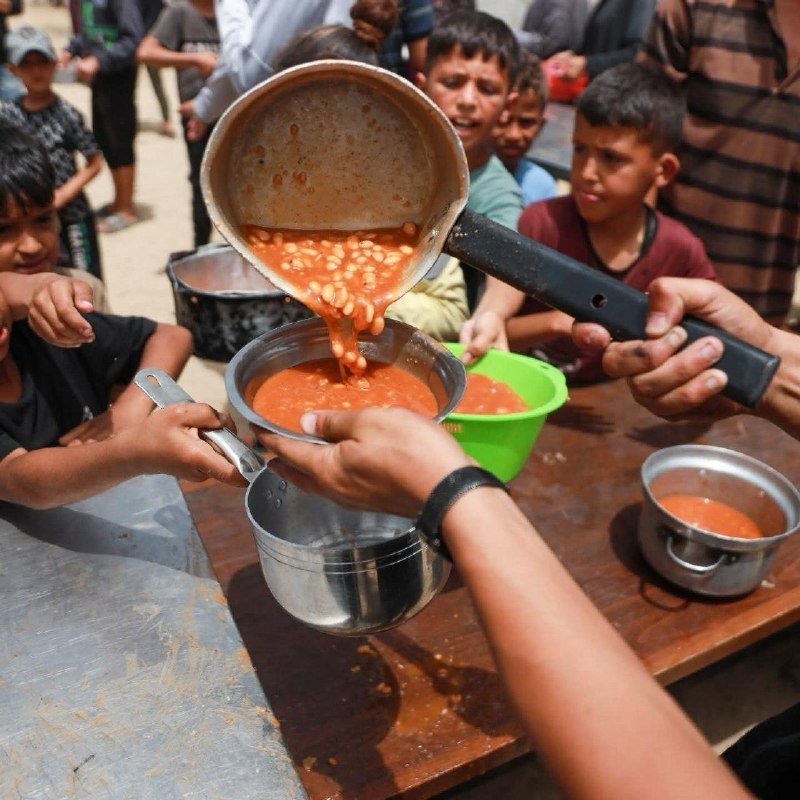 A person pours a red bean stew from a large pot into metal bowls and a green plastic bowl held by children gathered around a wooden table outdoors.