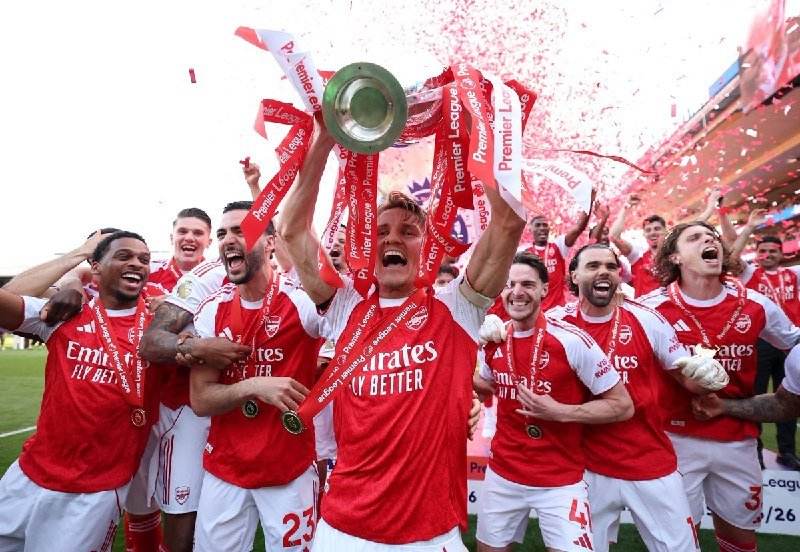 Soccer players in red "Emirates Fly Better" jerseys celebrate on a pitch, one hoisting a trophy with red ribbons as confetti falls around them.
