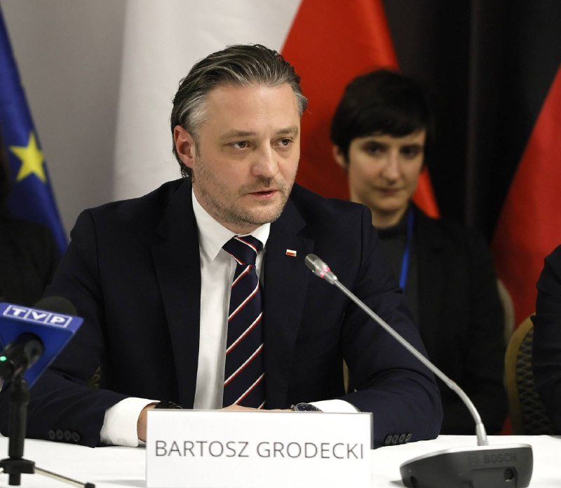 A man in a dark suit and striped tie sits at a table with a "BARTOSZ GRODECKI" nameplate, speaking into a microphone during a panel with flags behind him.