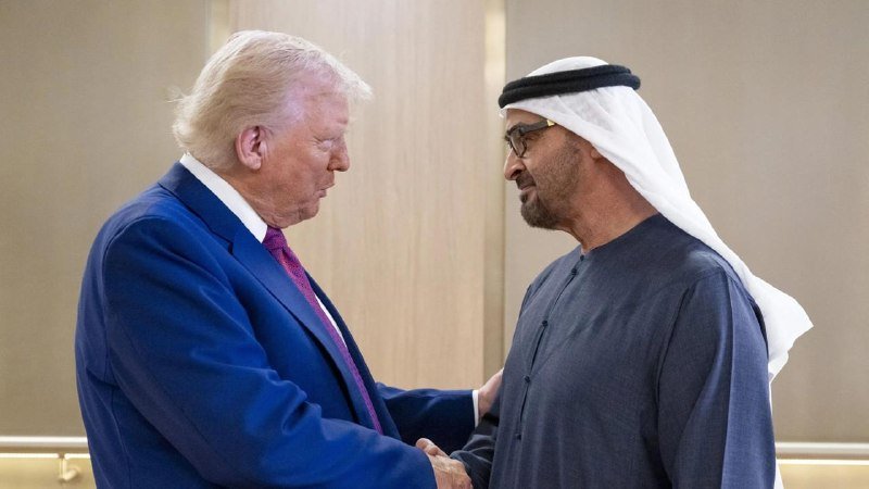 A man in a blue suit shakes hands with a man wearing a white headdress and dark traditional robe, standing face-to-face in an indoor setting.