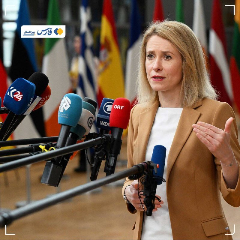 A blonde woman in a tan blazer speaks at a podium lined with international microphones, gesturing with one hand, while various national flags stand behind her.