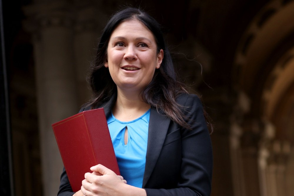 A smiling woman with long dark hair, wearing a black blazer over a blue top, holds a red folder while standing before an ornate stone archway.