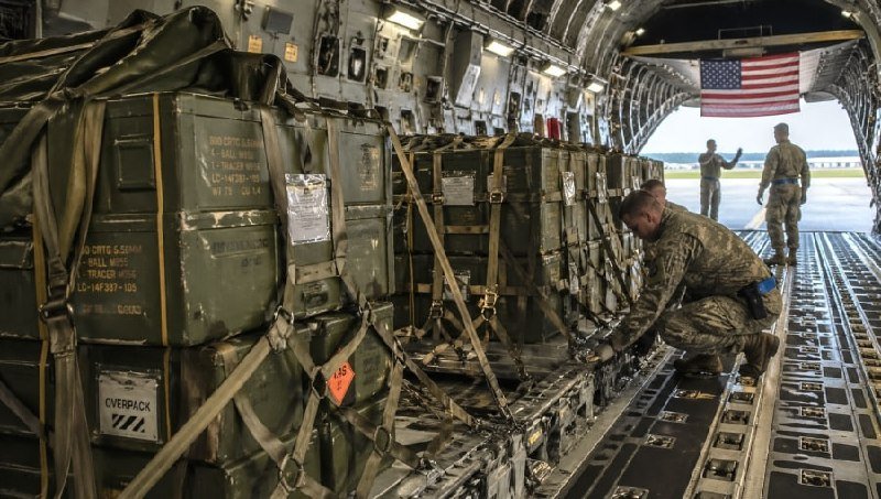 Soldiers in camouflage uniforms load or secure green military cargo pallets inside a transport aircraft with an American flag visible at the rear.