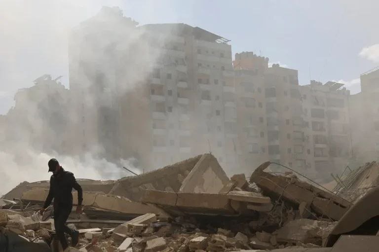 A man walks across a massive pile of concrete rubble as thick smoke rises from a collapsed multi-story building behind him.