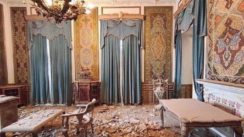 Debris litters the floor of a damaged, ornately decorated room with blue curtains, patterned wall panels, a chandelier, and scattered furniture.