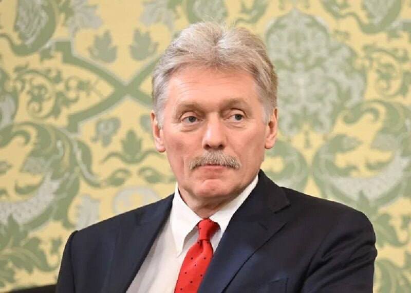 A gray-haired man with a mustache, wearing a dark suit and red tie, looks off to the side against a patterned green and beige background.