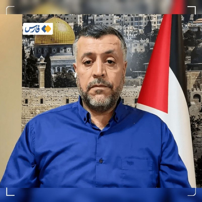 A bearded man in a blue shirt speaks before a backdrop showing the Dome of the Rock and a Palestinian flag, with a logo in the upper corner.