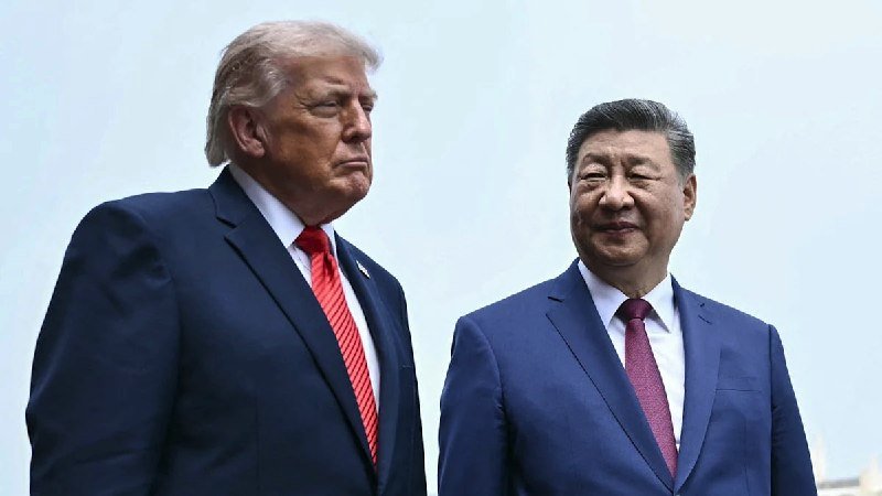 Two men in dark suits with red ties stand side by side outdoors against a pale sky, one with blond hair looking left, the other smiling toward the camera.