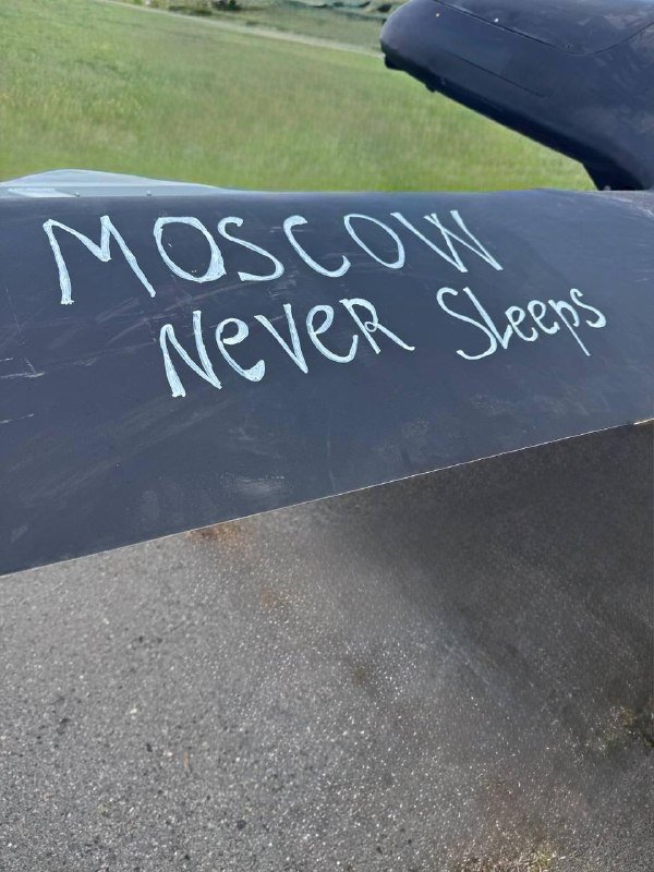 The phrase "MOSCOW Never Sleeps" is handwritten in white on a dark surface, set against a grassy background and paved ground.