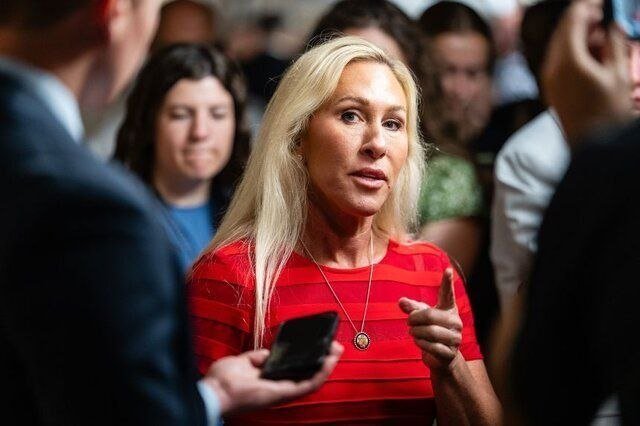 A blonde woman in a red top speaks while gesturing with her hand, surrounded by people, as someone holds a smartphone nearby.