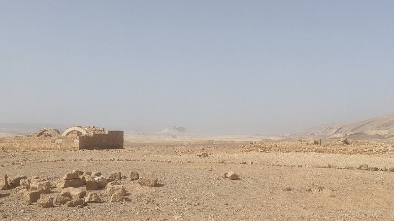 Stone ruins and scattered rubble sit in an arid desert landscape under a hazy sky, with barren mountains visible in the distance.