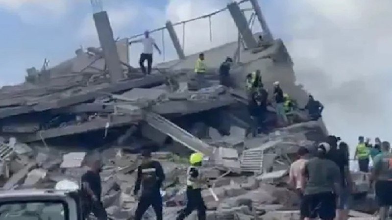 Rescue workers and bystanders climb through the rubble of a collapsed multi-story building, searching amid heavy debris under a clear sky.