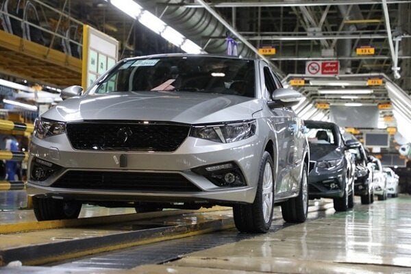 A silver sedan sits on an automotive assembly line in a factory, with additional vehicles lined up behind it under industrial lighting.