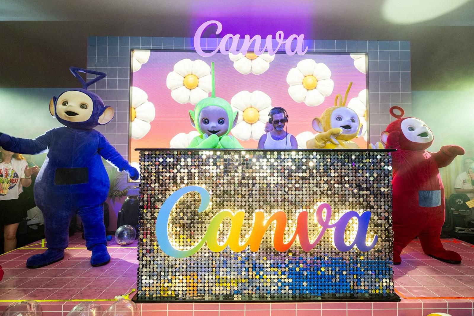 Costumed Teletubby mascots pose beside a DJ booth displaying the "Canva" logo at a branded event.