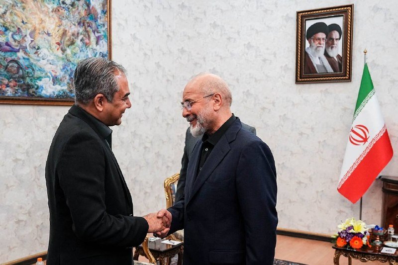 Two men in dark suits shake hands and smile in a formal room featuring an Iranian flag, a framed portrait of two clerics, and a colorful painting.