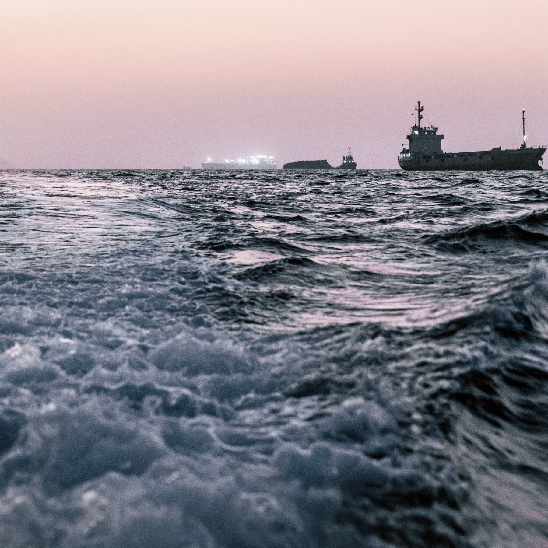 Cargo ships sit on choppy open water under a pale pink and gray dawn or dusk sky.
