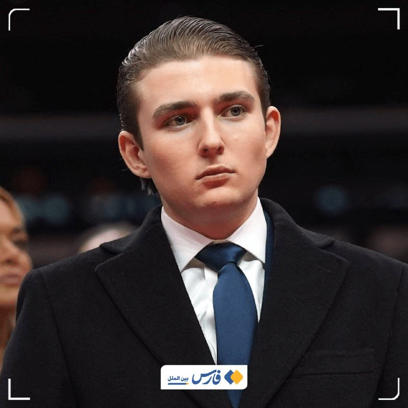 A young man with slicked-back dark hair wearing a black coat, white shirt, and blue tie stands against a dark background.